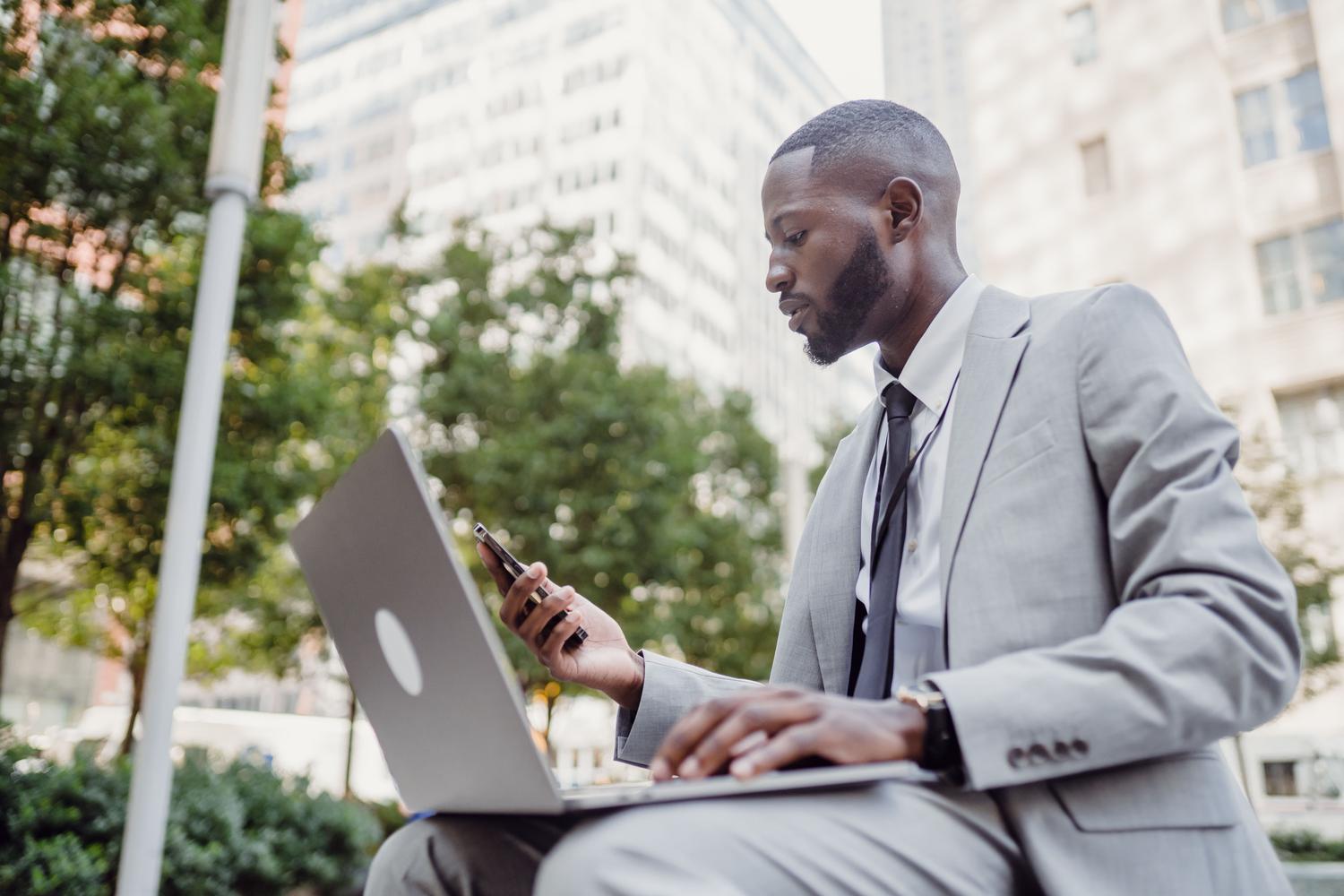 Hombre de negocios utilizando simultáneamente un portátil y un smartphone en un entorno urbano, representando la gestión móvil de un negocio.