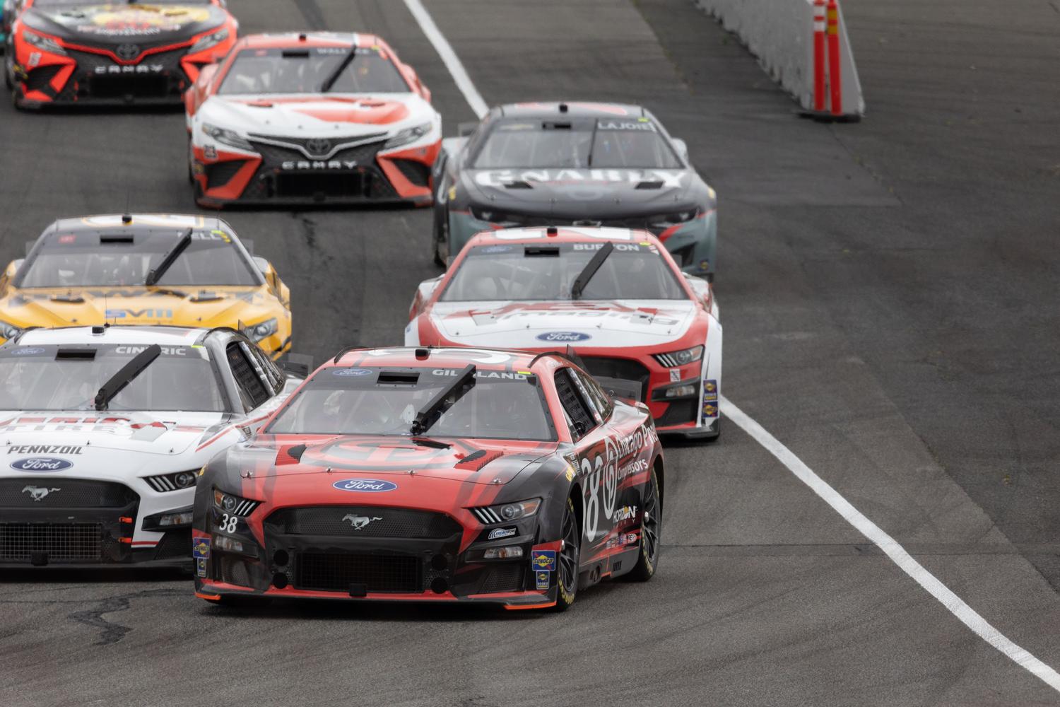 Pack de coches NASCAR compitiendo a alta velocidad en un óvalo, representando la intensidad de las carreras en NASCAR 26.