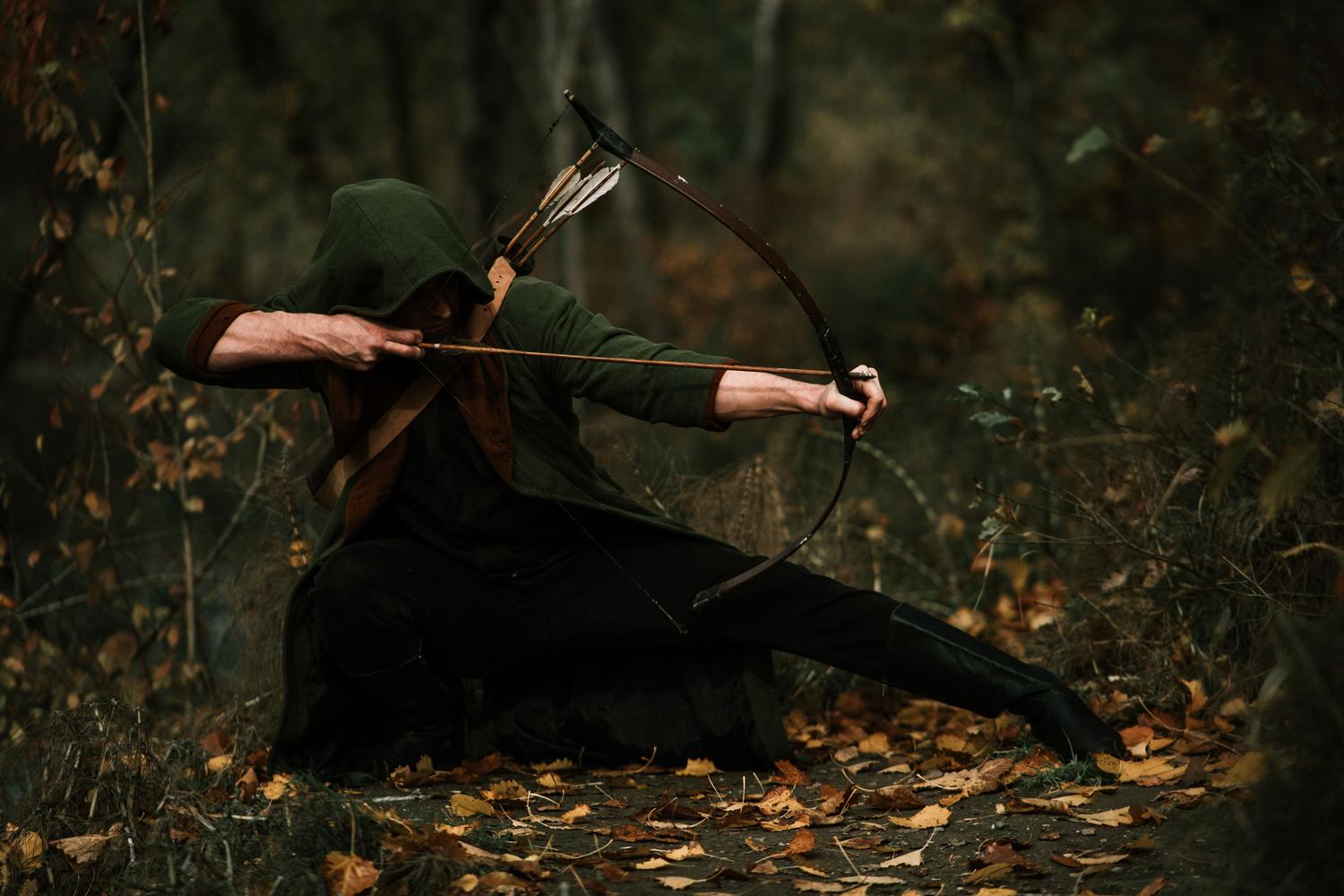 Arquero medieval con capucha verde acechando en el bosque, ilustrando el arquetipo de juego del Arquero