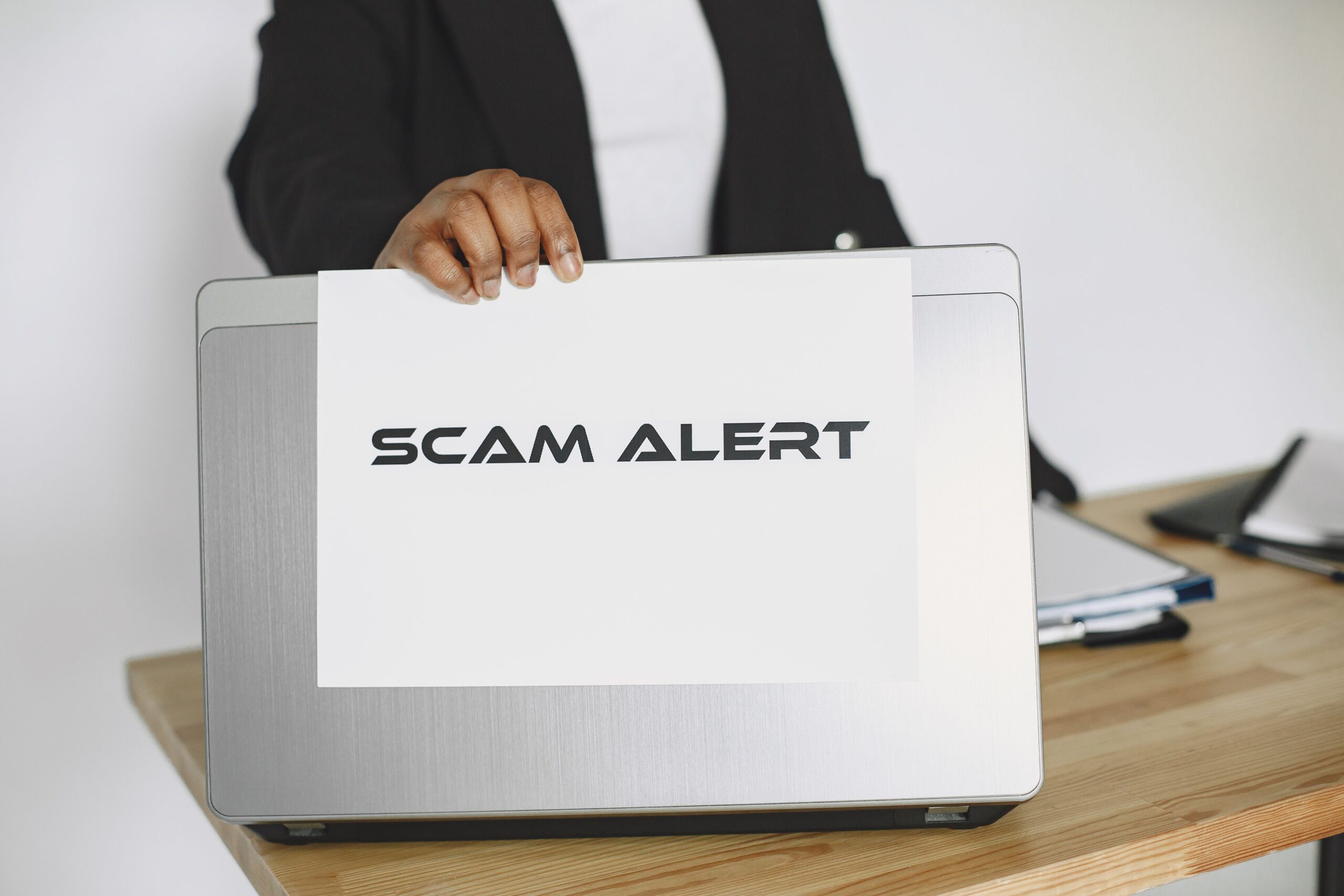 Persona sosteniendo un cartel de alerta de estafa (SCAM ALERT) sobre un ordenador portátil.