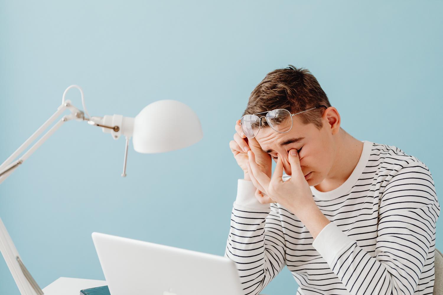 Joven cansado frotándose la nariz y los ojos mientras trabaja con un portátil bajo la luz de una lámpara.