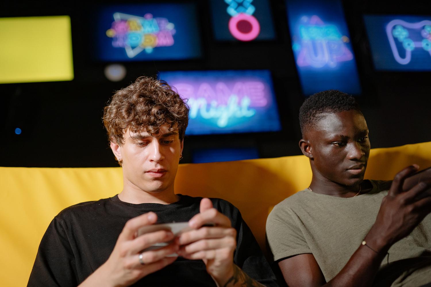 Dos amigos concentrados jugando con sus teléfonos móviles en un interior, ejemplificando la naturaleza social y competitiva de los servicios de juegos.