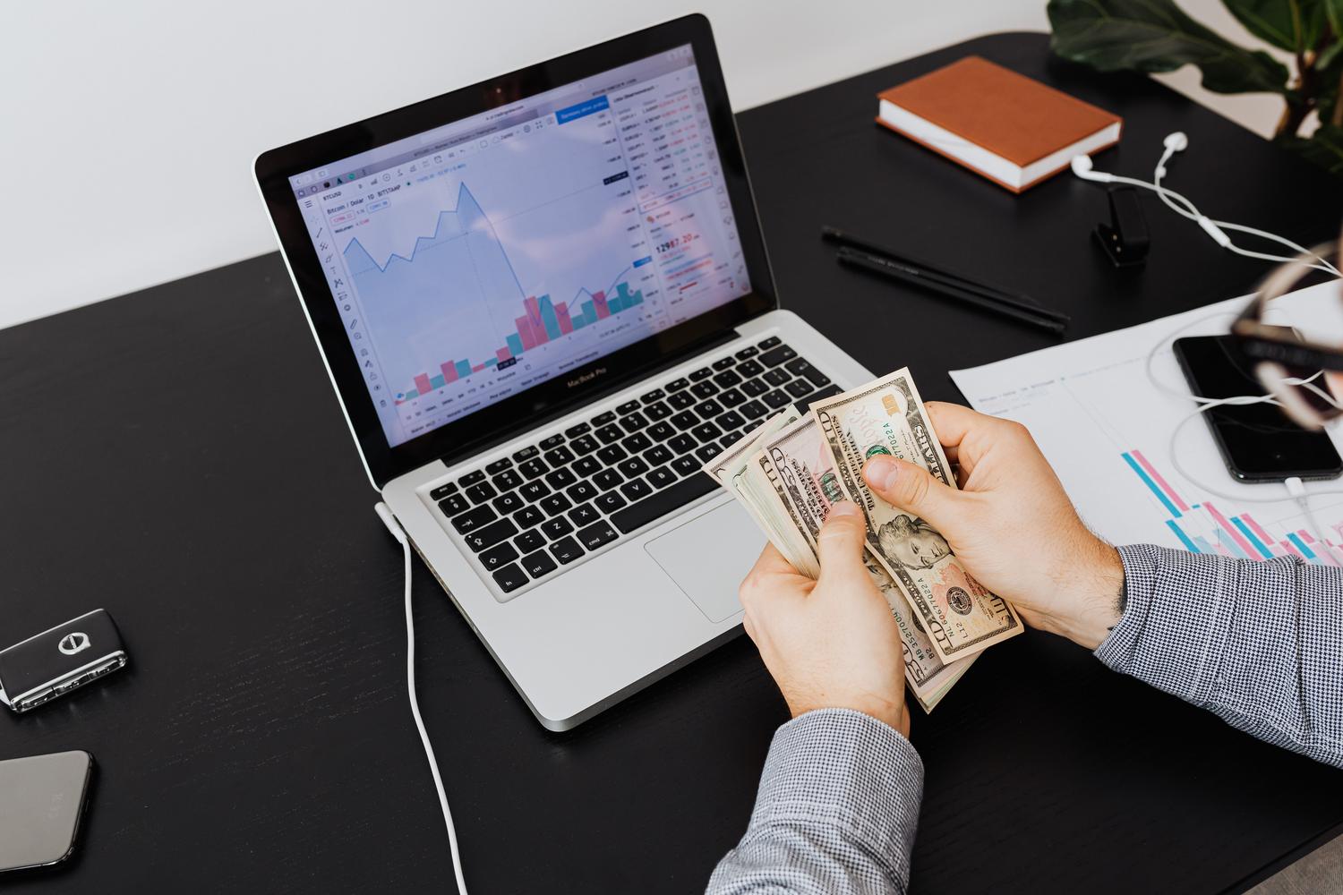 Persona analizando gráficos financieros en un portátil mientras sostiene billetes de dólar, simbolizando el éxito financiero en el marketing de afiliados.