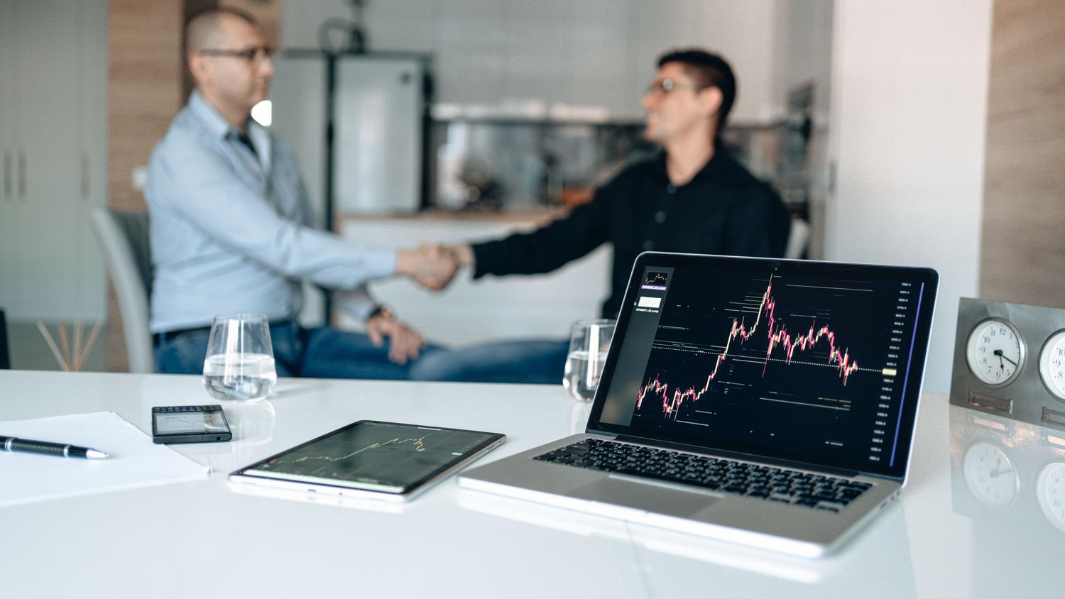 Dos profesionales dándose la mano en una oficina con gráficos financieros de fondo, simbolizando una alianza estratégica entre empresa y afiliado.
