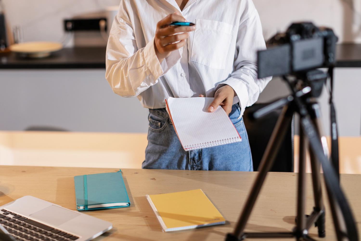 Creadora de contenido preparando un guion con su portátil y cámara en un trípode, representando la labor del blogger o influencer en la promoción de productos.