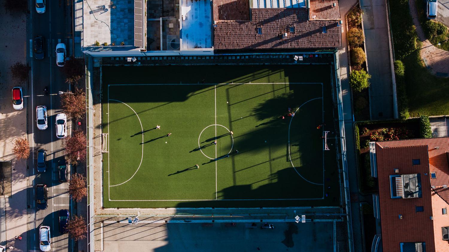 Vista aérea de personas jugando al fútbol en una cancha urbana rodeada de edificios, representando la escala social de The Grounds.