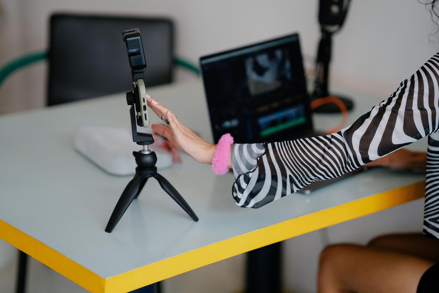 Creadora de contenido digital utilizando un ordenador portátil y un trípode con smartphone para la edición de vídeo y diseño.