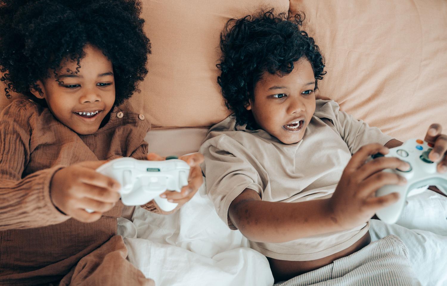 Dos niños pequeños jugando videojuegos cómodamente en su cama, transmitiendo la facilidad de acceso al juego.