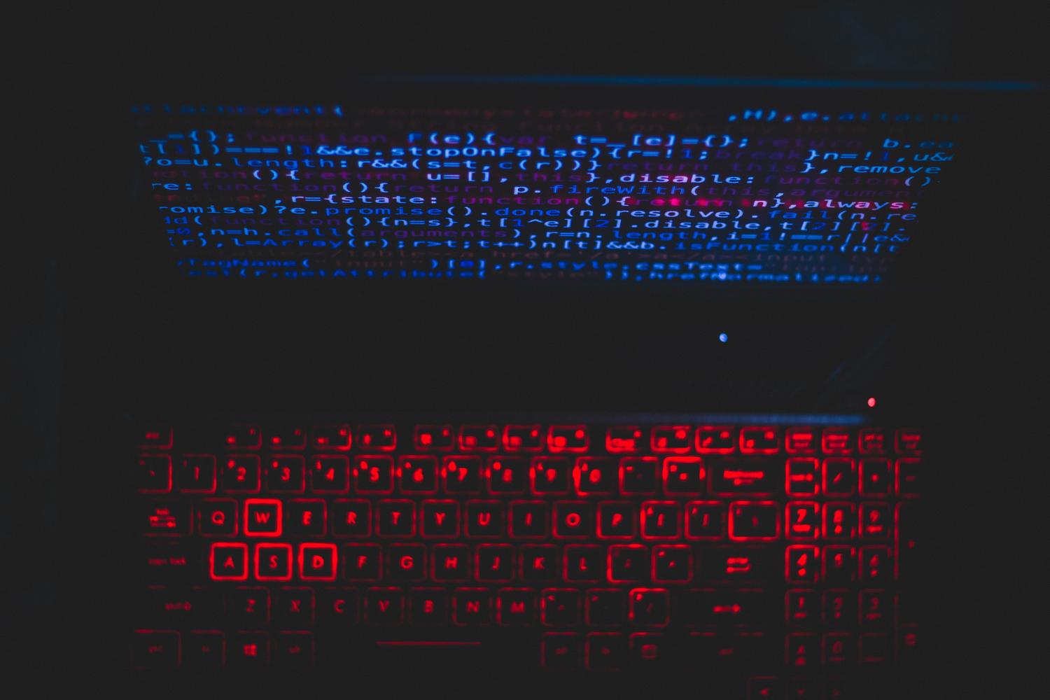 Teclado retroiluminado en rojo y código de programación en pantalla, ilustrando los riesgos de ciberataques y fugas de datos.
