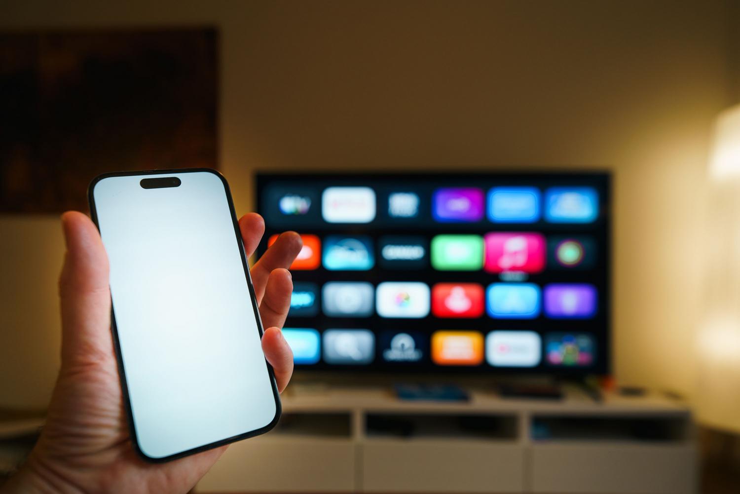 Smartphone en primer plano con Smart TV difuminada al fondo mostrando un menú de aplicaciones, simbolizando la compatibilidad entre dispositivos móviles y televisores.