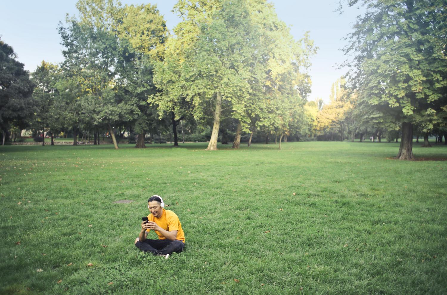 Hombre sentado en el césped de un parque usando su smartphone, representando la búsqueda de Pokémon en el entorno natural