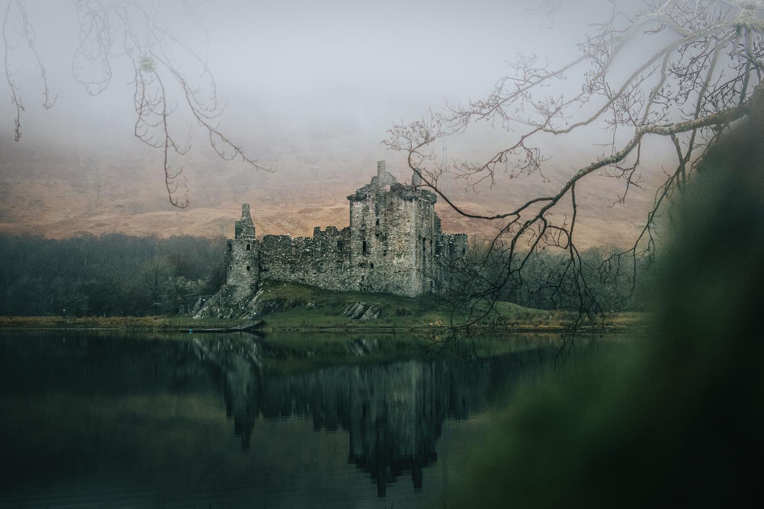 Castillo medieval envuelto en niebla reflejado en un lago oscuro, evocando la atmósfera melancólica de las Tierras Intermedias.