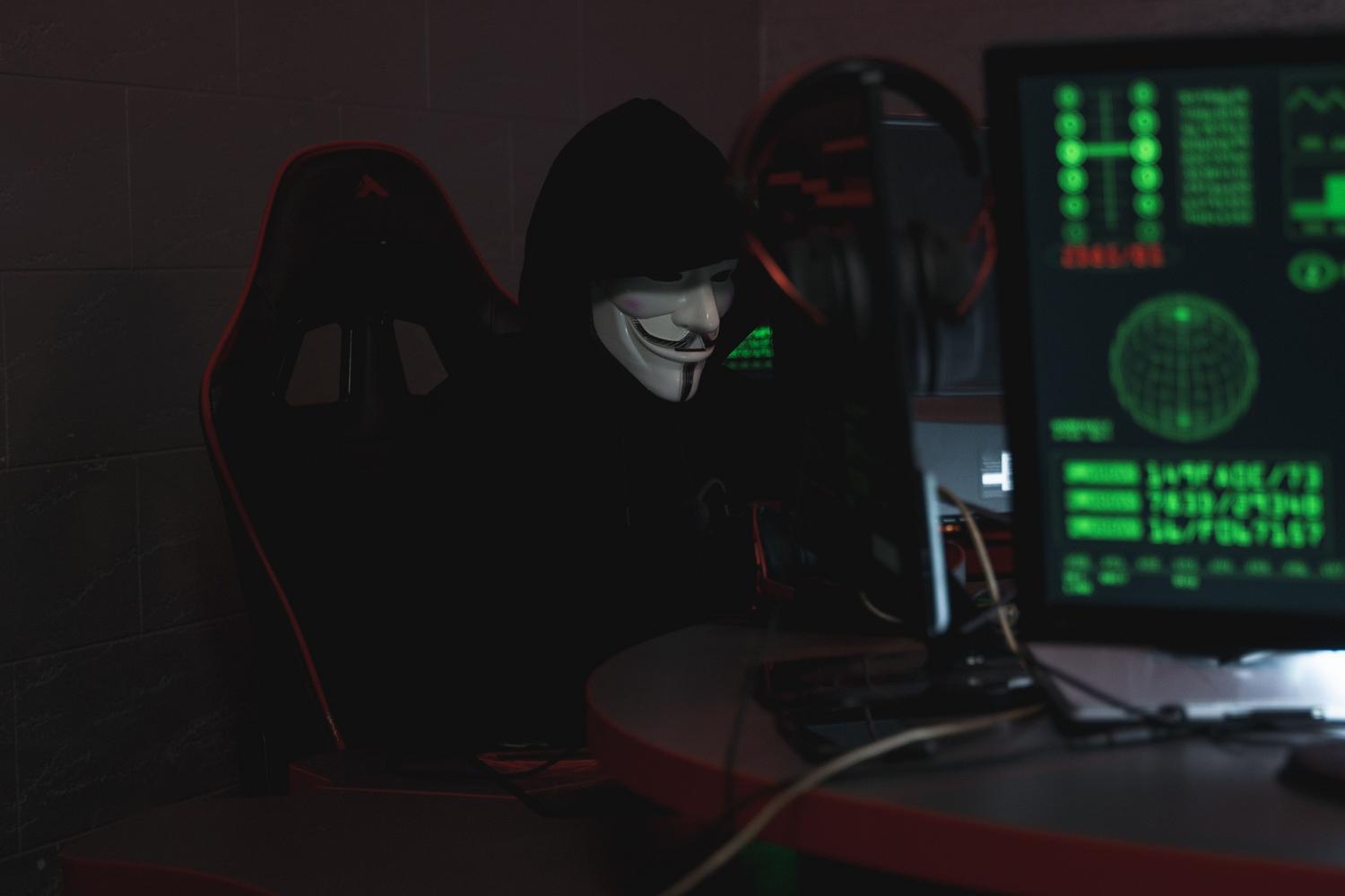 Persona con máscara de Guy Fawkes frente a un ordenador en una habitación oscura, representando el anonimato de la Dark Web.