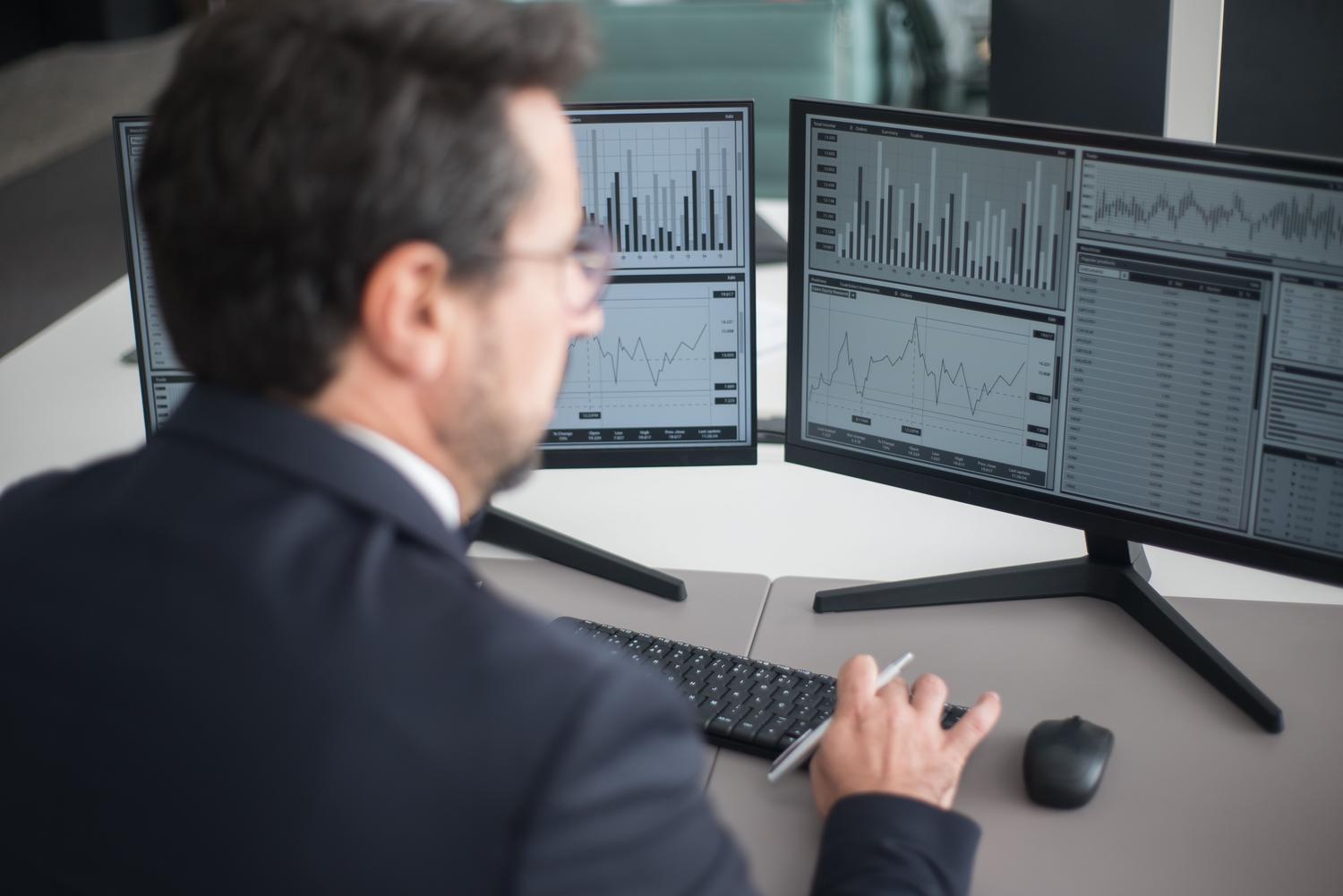 Hombre de negocios analizando gráficos financieros complejos en dos monitores, representando el trabajo con hojas de cálculo avanzadas.