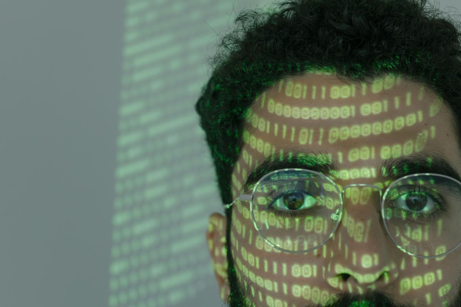 Primer plano de un experto en ciberseguridad con código binario proyectado en su rostro y gafas, simbolizando la simbiosis entre el humano y la máquina.