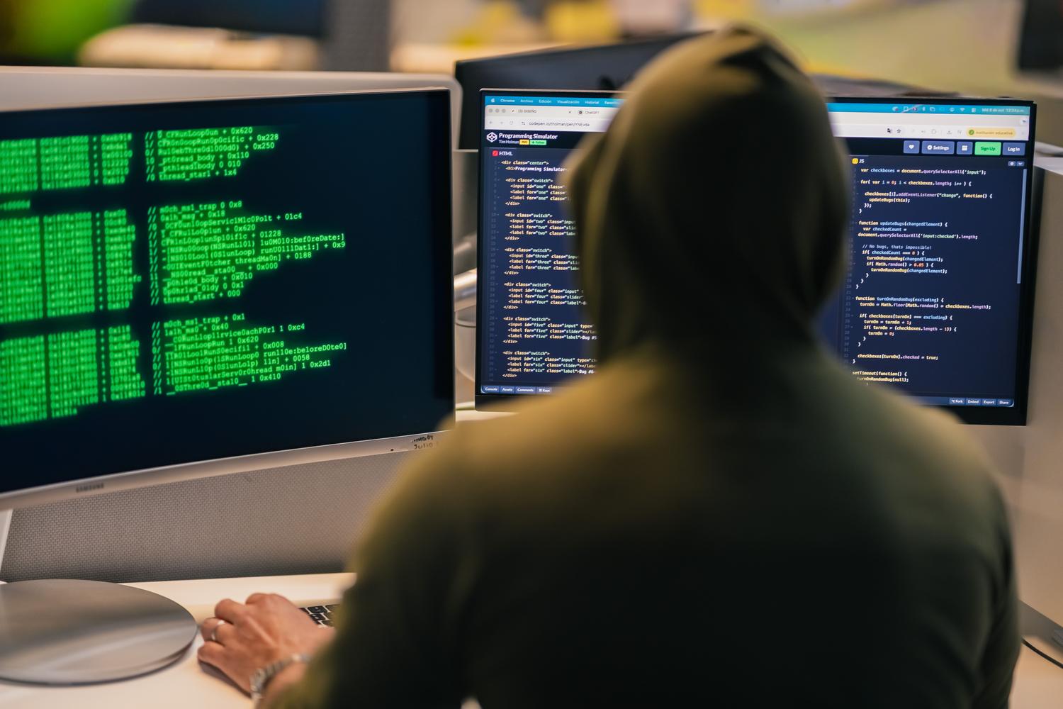 Persona con capucha trabajando con múltiples monitores en un ambiente oscuro, ilustrando la amenaza constante y la sofisticación del hacking.