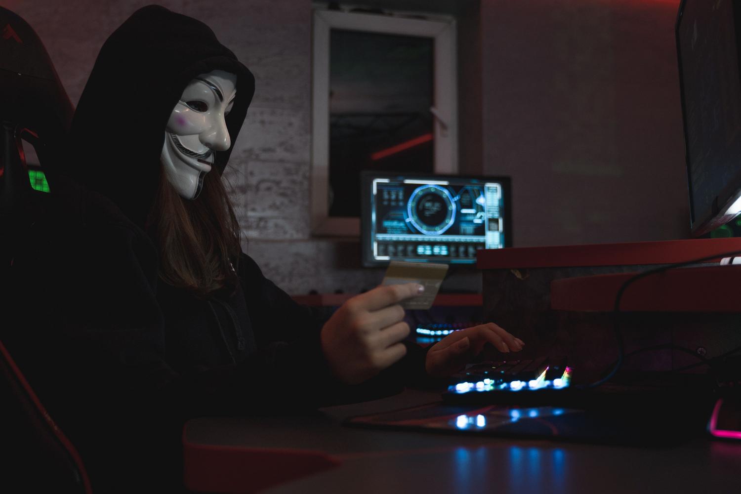 Hacker con máscara de Guy Fawkes sosteniendo una tarjeta de crédito frente a un ordenador, representando el fraude financiero y el robo de datos.
