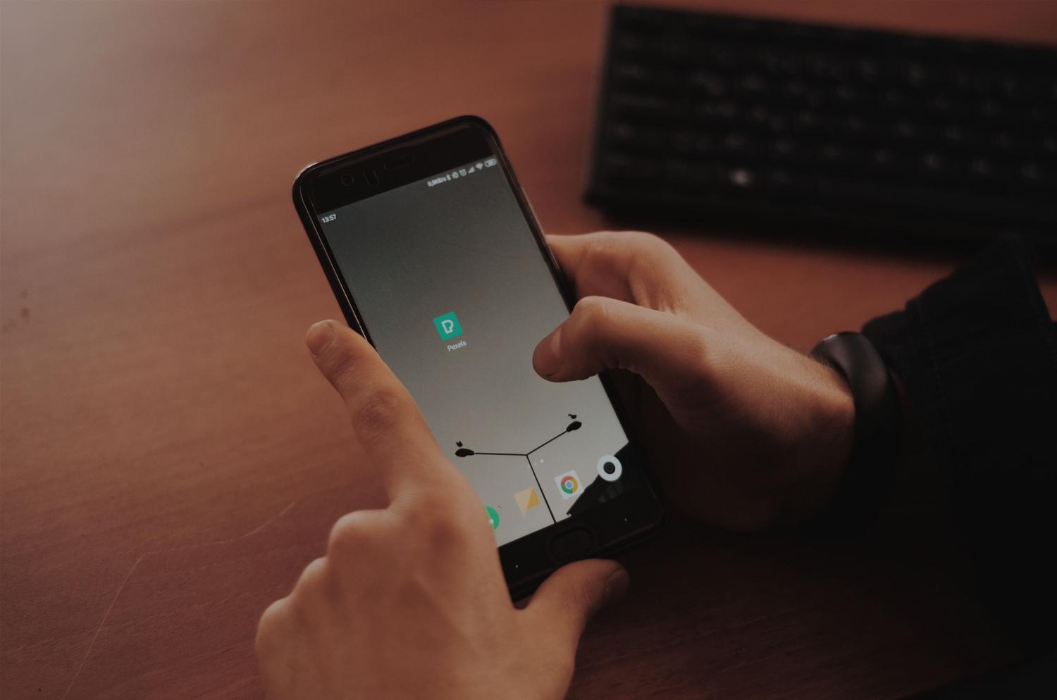 Primer plano de manos interactuando con un smartphone sobre un escritorio de madera con un teclado al fondo, ideal para representar el uso de WhatsApp Business o Web.