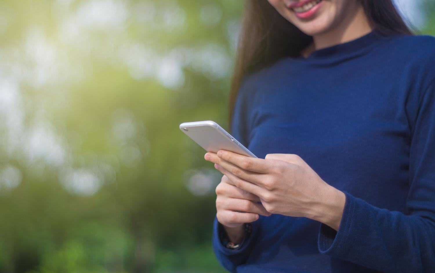 Mujer sonriente usando un smartphone blanco en un parque soleado con fondo verde desenfocado, representando la tranquilidad de tener la comunicación organizada.