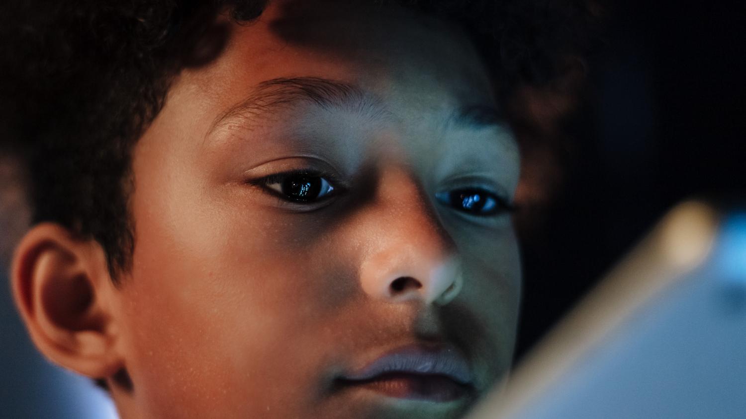 Retrato de un niño absorto en la luz azul de una pantalla en la oscuridad, simbolizando la vulnerabilidad y los riesgos del entorno digital.