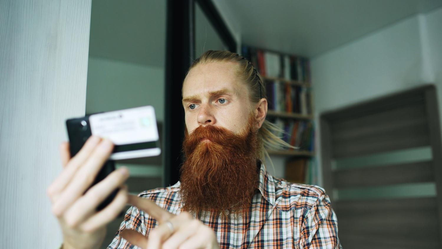 Hombre utilizando un smartphone y una tarjeta de crédito en un entorno doméstico, ilustrando el uso de tarjetas bancarias como filtro de acceso rápido para adultos.