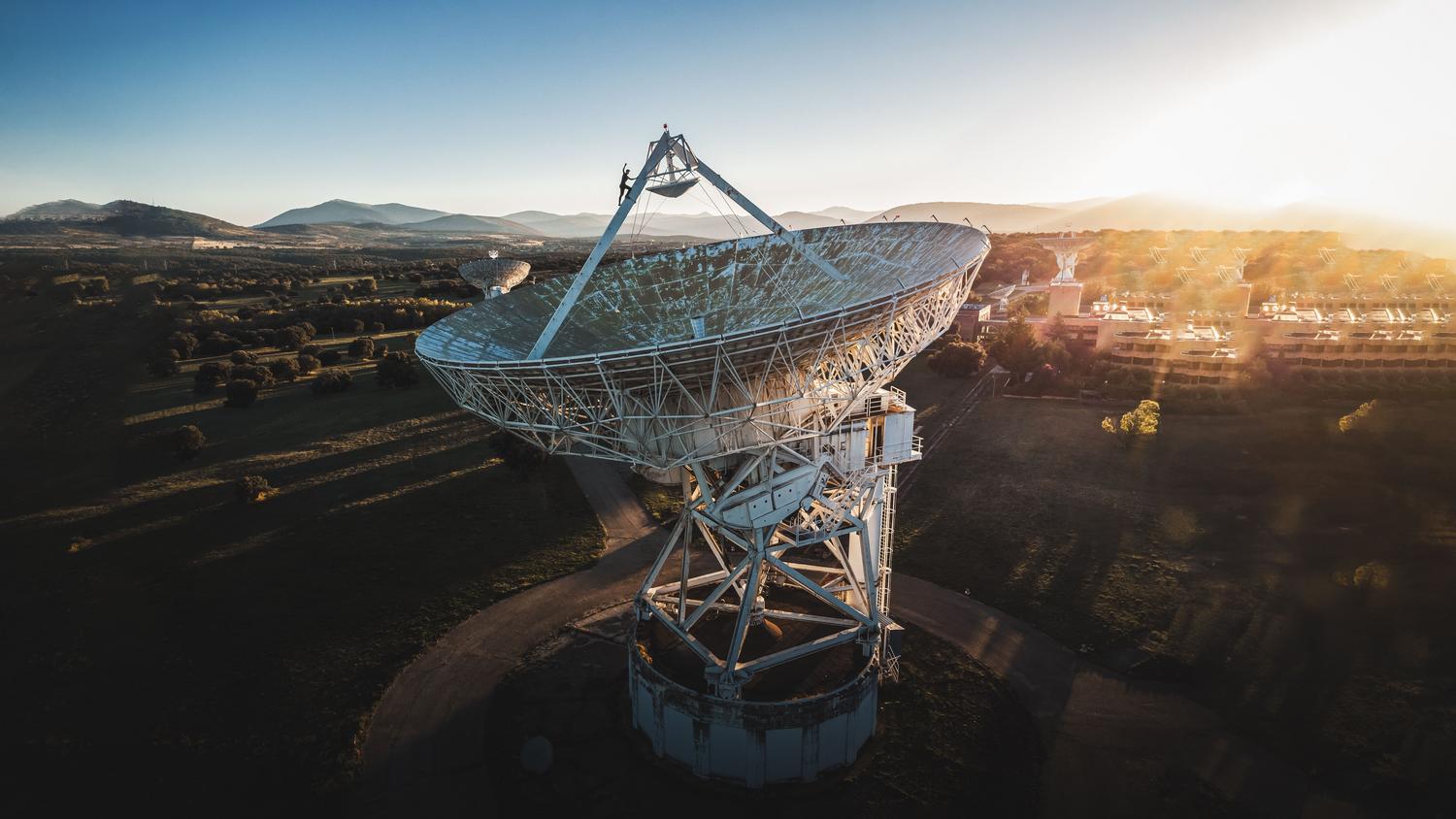 Vista aérea de una gran antena parabólica en un paisaje rural durante el amanecer, representando las estaciones terrestres de conexión.