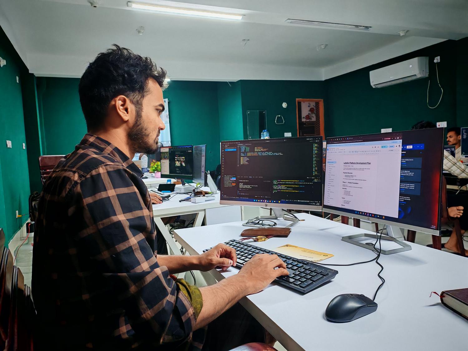 Software developer working on code at a dual monitor setup in a modern office environment.