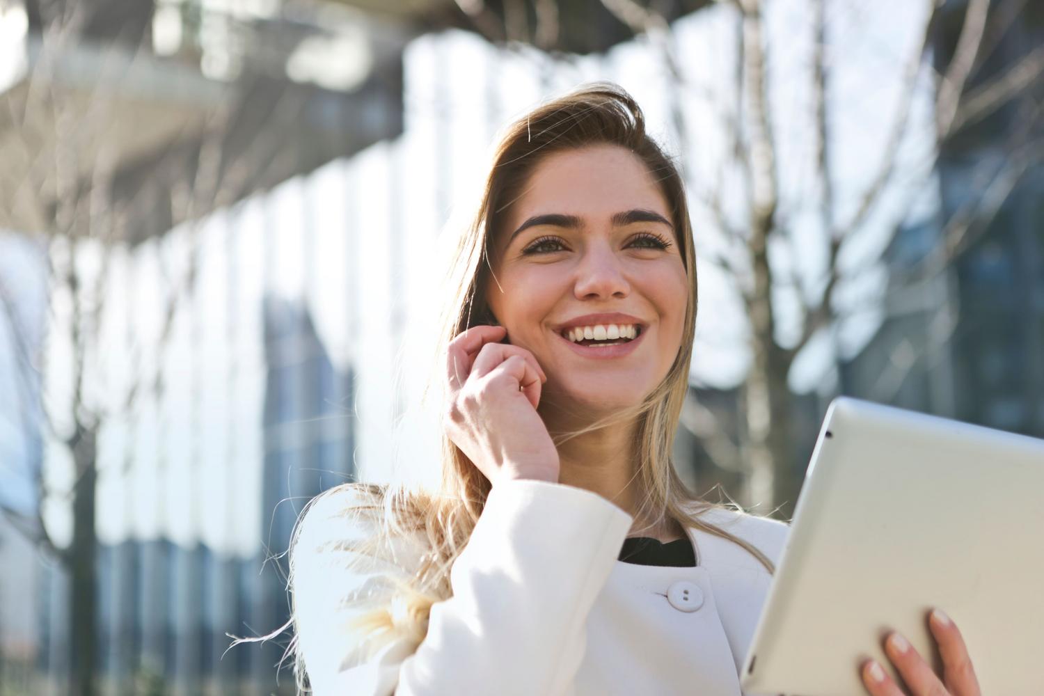 Mujer de negocios utilizando una tablet y un teléfono móvil en un entorno urbano, representando la movilidad y el acceso a la información empresarial desde cualquier lugar.