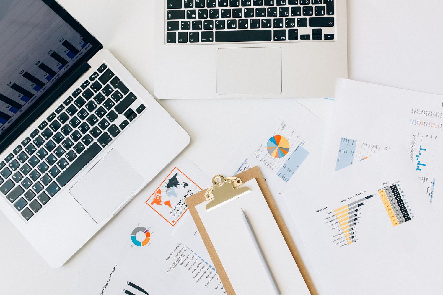 Vista cenital de un escritorio con portátiles y diversos informes con gráficos, ideal para ilustrar la gestión de datos complejos y la conversión de formatos.