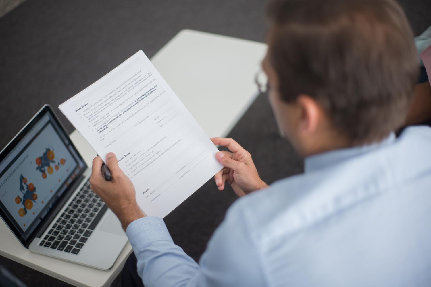 Profesional revisando un documento impreso comparándolo con datos en un portátil, reflejando la fase de revisión de informes corporativos.