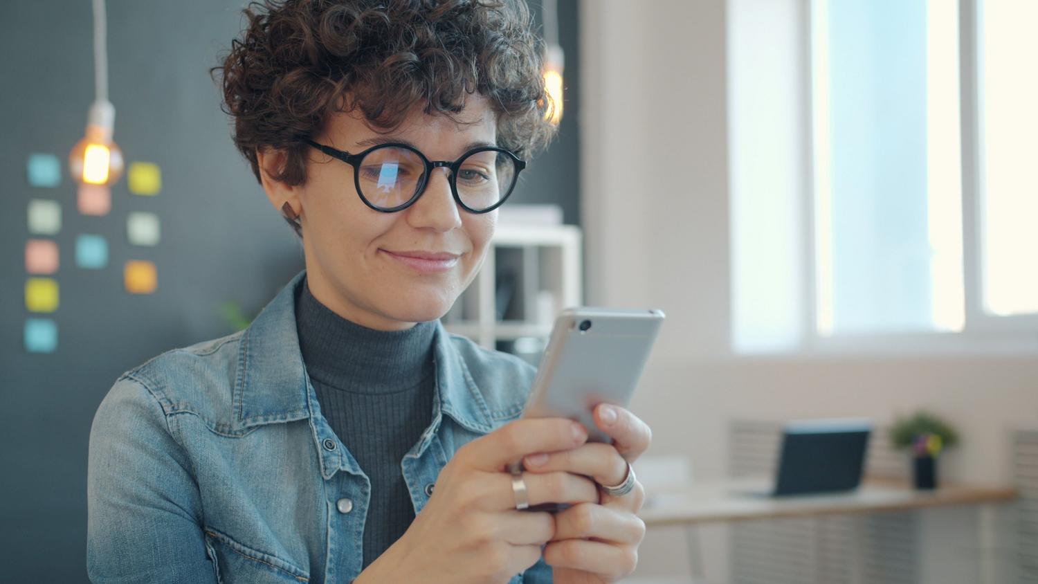 Mujer en una oficina moderna usando su teléfono móvil con expresión satisfecha, ilustrando la organización de recordatorios en la rutina diaria.