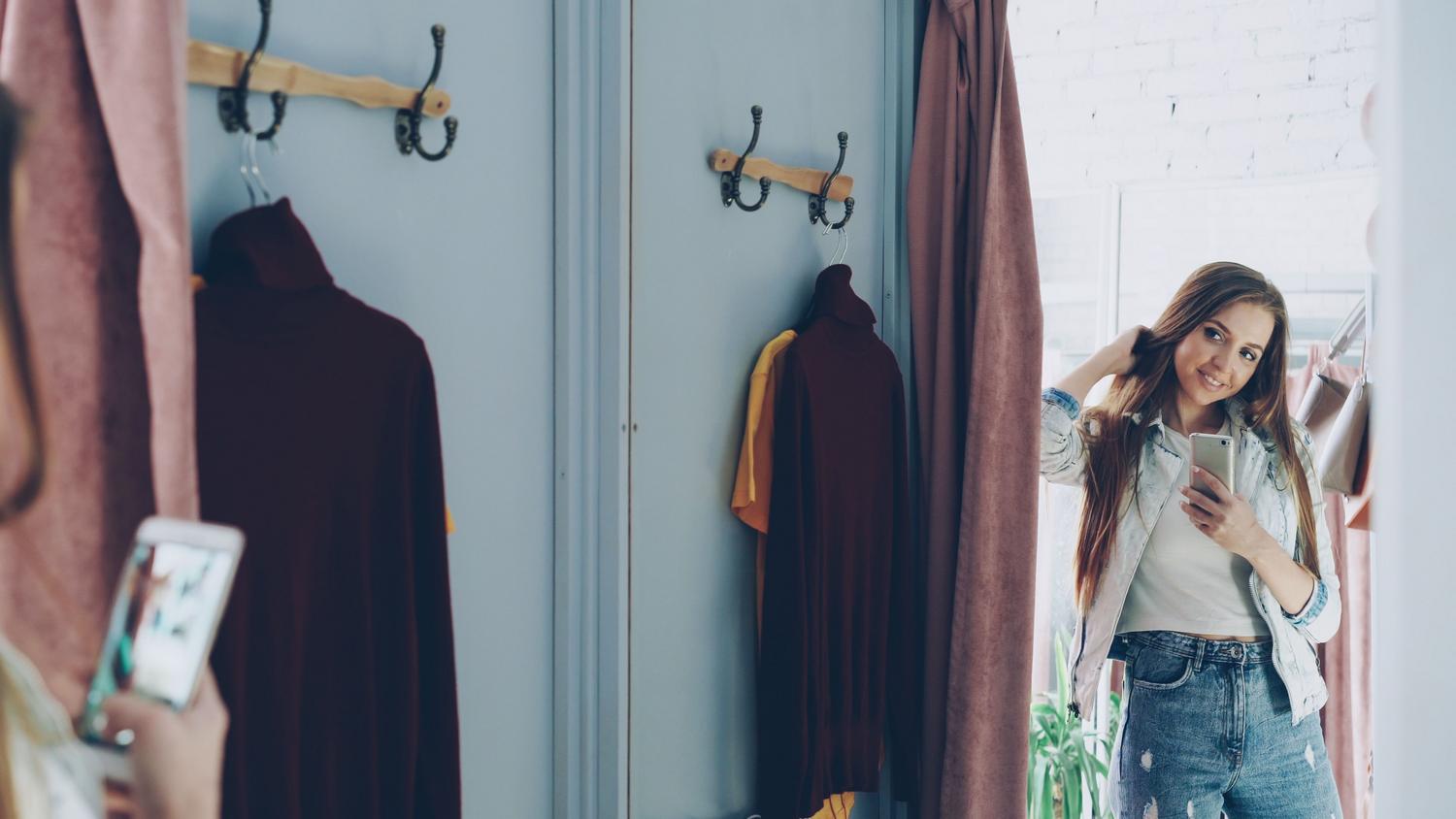 Young woman taking a selfie in a physical store fitting room, illustrating the traditional shopping process that virtual try-on tools aim to simplify.