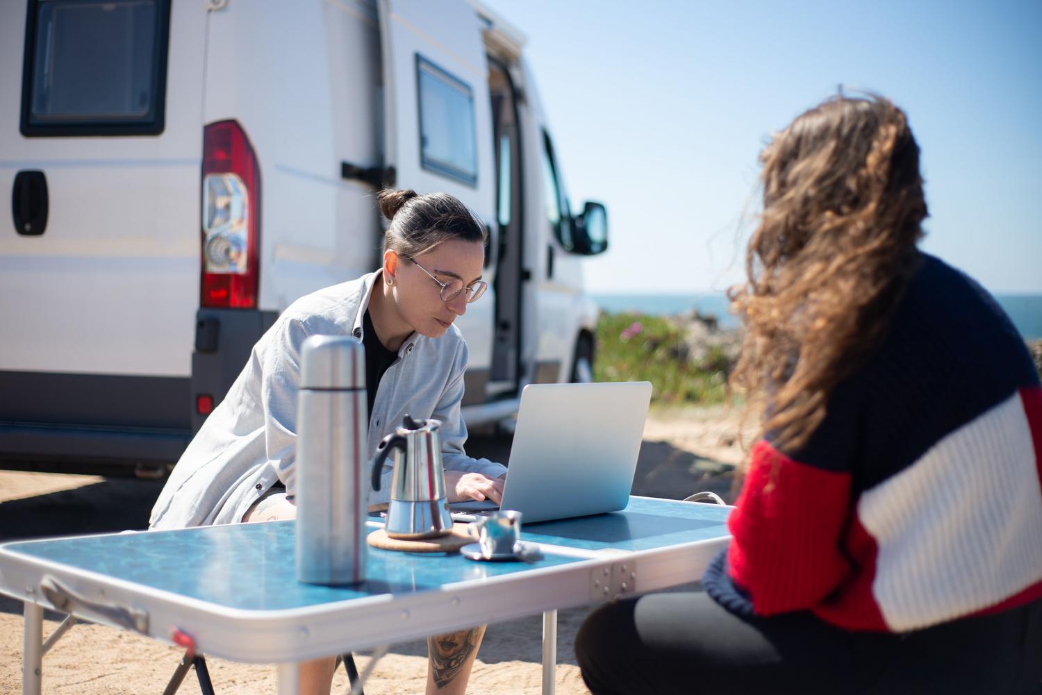 Nómadas digitales trabajando con portátil junto a una furgoneta camper en la playa